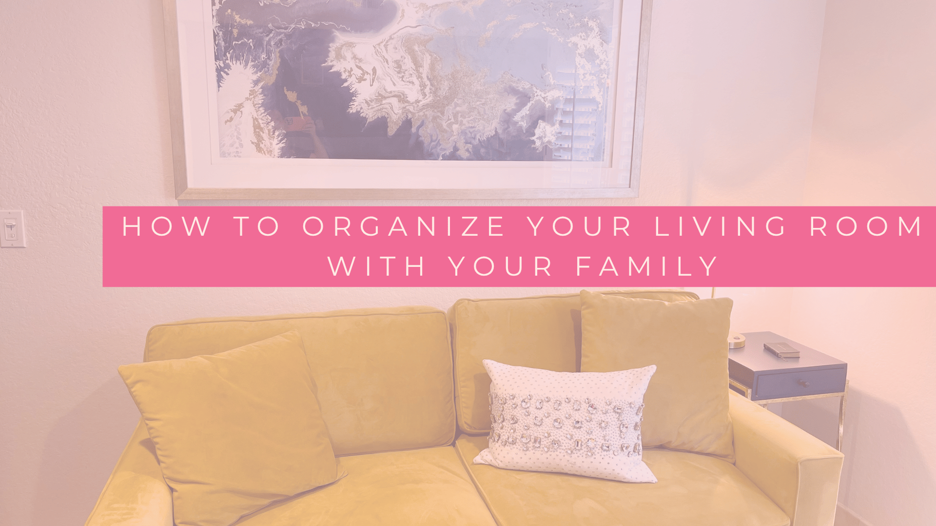 A warm, styled living room with a gold velvet sofa and a jeweled accent pillow beneath abstract blue-and-gold art, with text reading How to Organize Your Living Room With Your Family.
